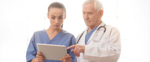 Imagem de um homem grisalho com jaleco apontando para um tablet que está na mão de uma mulher mais jovem que parece ser uma enfermeira.